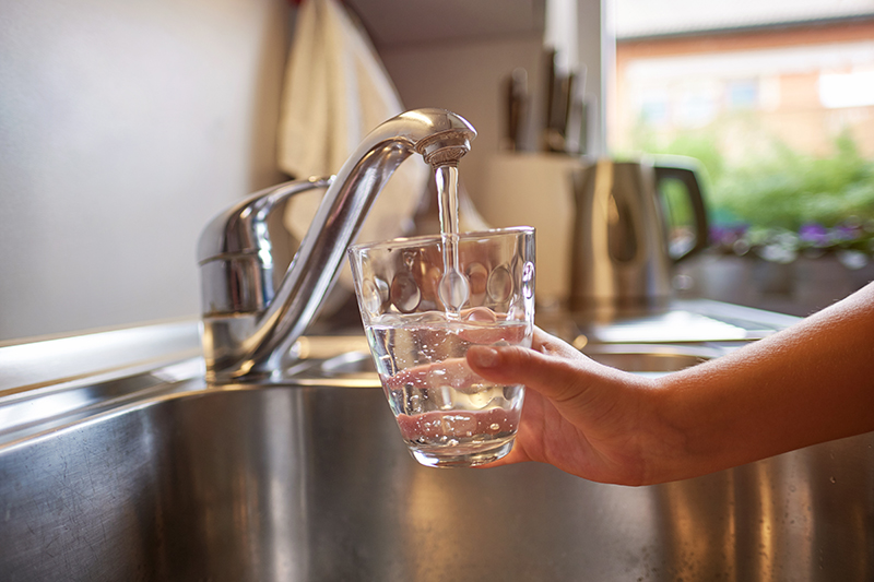 Close up of a hand filling a clear glass of water from a kitchen sink faucet.