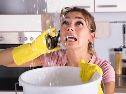 Concerned woman on the phone holding a bucket that water is streaming into from above.